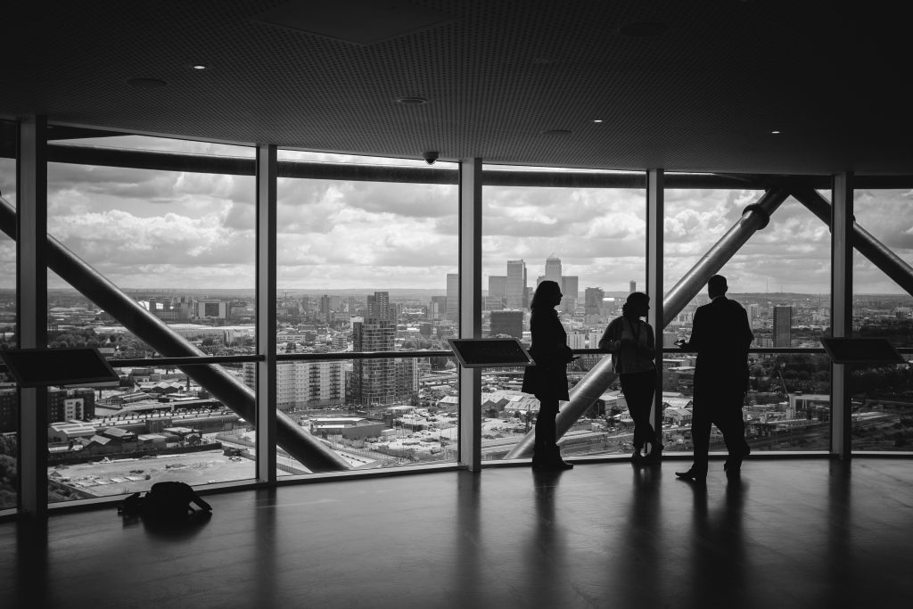 Silhouettes of business professionals overlooking a city skyline from a high-rise office, representing The On-Demand CIO/CSO and scalable, expert advice at a flexible cost.